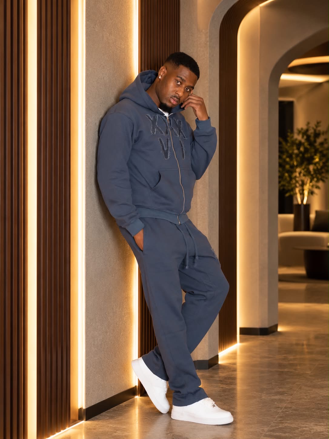 A man wearing a matching slate blue zip-up hoodie and jogger pants set with white sneakers, leaning against a modern wall with warm vertical light strips in an indoor hallway.